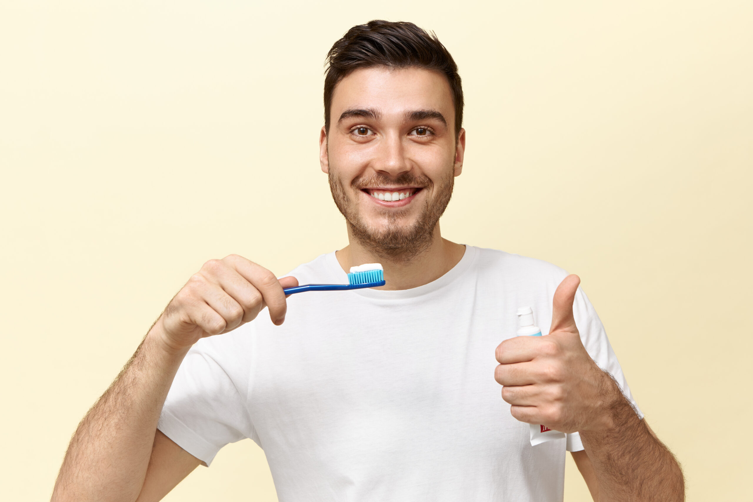 Happy energetic young European guy with stubble holding tooth brush with whitening paste and showing thumbs up gesture being in good mood. Dental care, oral cavity hygiene and healthy teeth concept